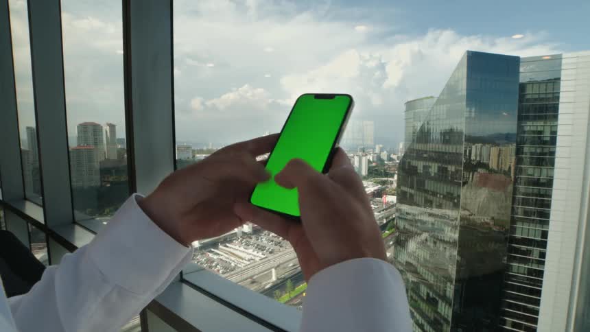 A businessman's hands scroll on a smartphone with a green screen in front of a window overlooking a modern city skyline