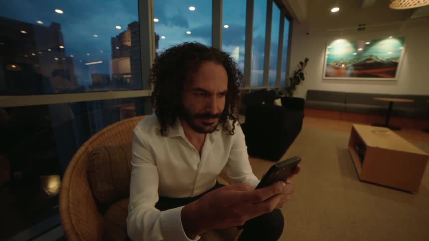 A focused man with curly hair sits in a modern lounge area at dusk, scrolling through his smartphone