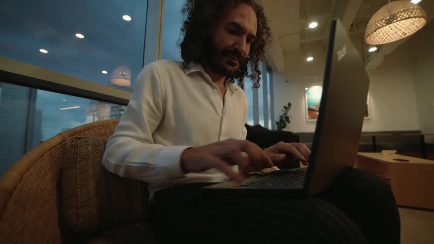 A focused professional man with curly hair works late on his laptop in a modern high-rise office lounge as the camera slowly pushes in