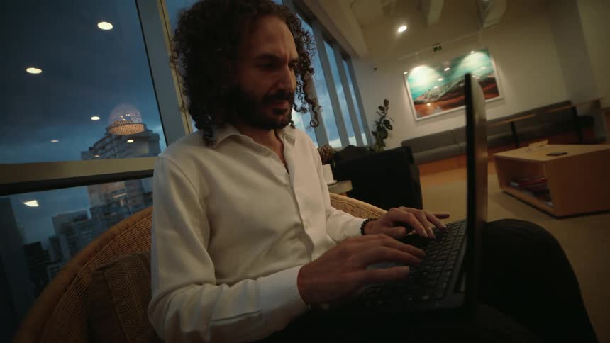 Focused professional man with curly hair works on his laptop in a modern office lounge overlooking the city at dusk