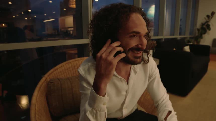 A cheerful man with curly hair has a pleasant phone conversation while sitting in a chair in a modern apartment at night