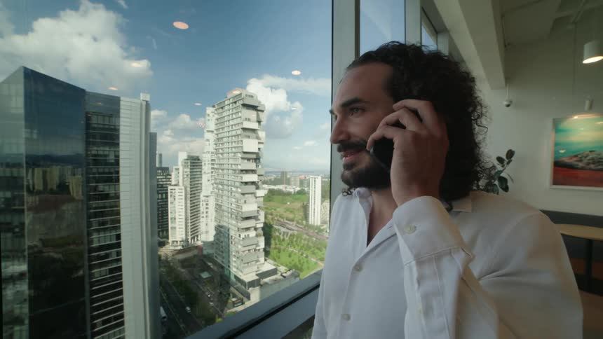 A smiling businessman with curly hair talks on his phone while looking out the window of a modern high-rise office at the city skyline