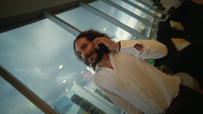 A handsome man with long curly hair answers a phone call in his modern high-rise apartment overlooking the city