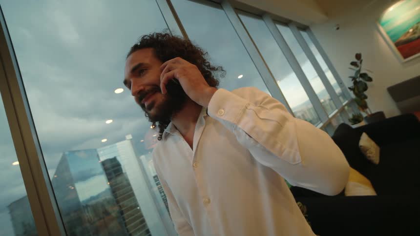 A smiling businessman with long curly hair makes a phone call while standing in a modern office with a large window overlooking the cityscape