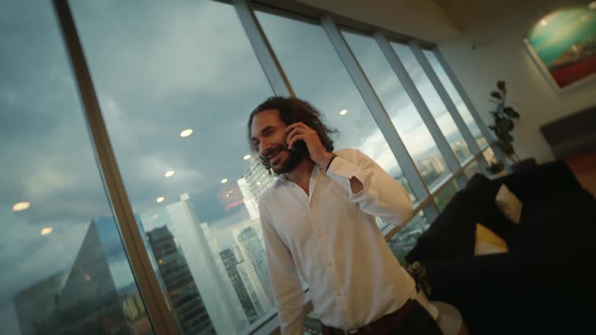A businessman with long curly hair smiles while talking on the phone in a modern high-rise office with a city view
