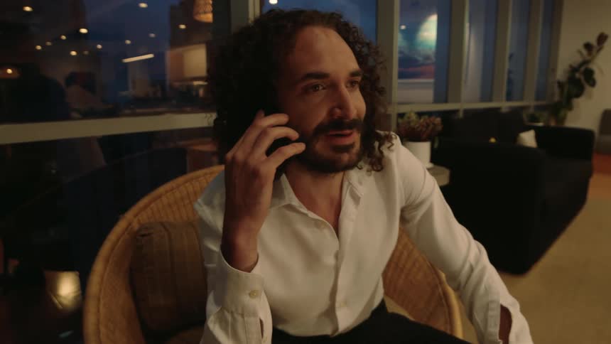 A man with curly hair and a white shirt sits in a wicker chair while talking on his phone in a modern living room at night