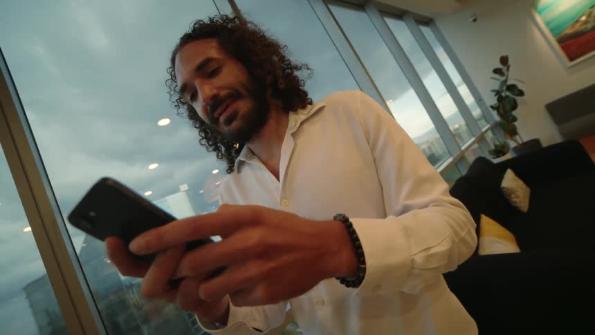 A curly-haired businessman smiles while using his smartphone in a modern high-rise office with a cityscape view
