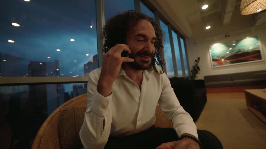 A smiling man with curly hair talks on his smartphone while sitting in a modern high-rise office lounge in the evening