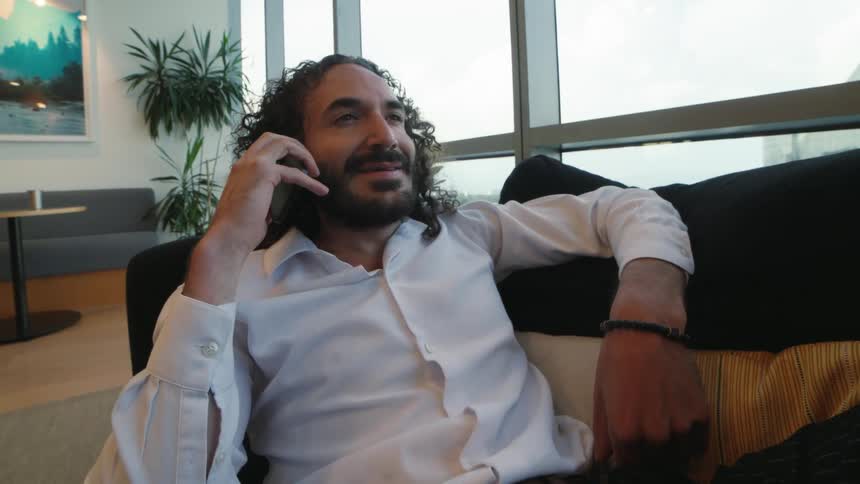 A relaxed man with curly hair and a beard answers his phone while sitting on a couch in a modern high-rise