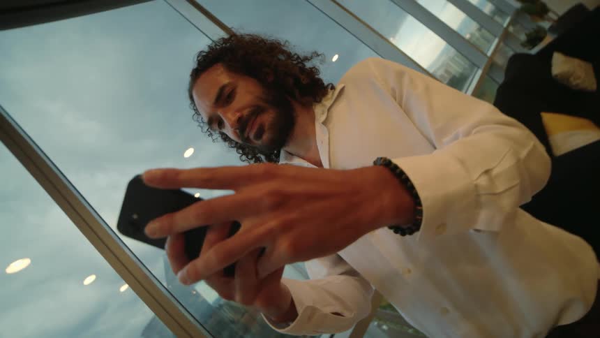 A man with curly hair stands next to a window in a high-rise building as he smiles and scrolls on his smartphone