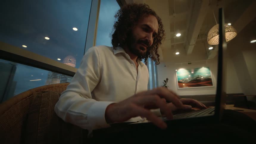 A focused professional with curly hair works on his laptop in a modern office with a city skyline view at dusk