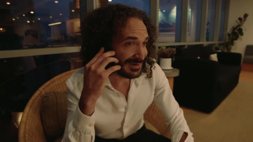 A smiling man with long curly hair has a pleasant conversation on his smartphone while sitting indoors at night
