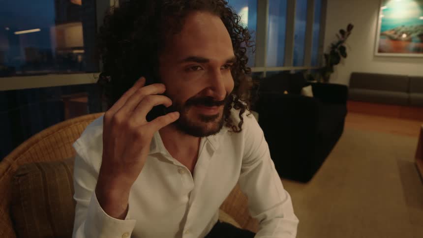 A professional man with a beard and curly hair engages in a friendly phone call while sitting in his modern living room at dusk