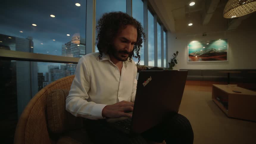 A focused professional man with curly hair works diligently on his laptop in a modern high-rise office as dusk settles over the city