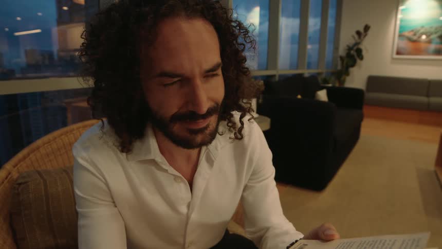 A relaxed man with long curly hair enjoys reading a book in his modern high-rise apartment at night