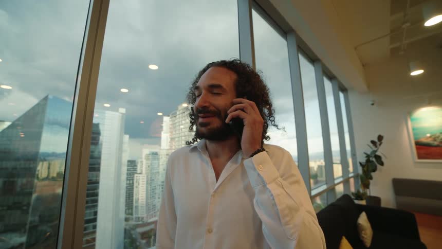 A successful businessman with long curly hair smiles while talking on the phone in his modern high-rise office overlooking the city