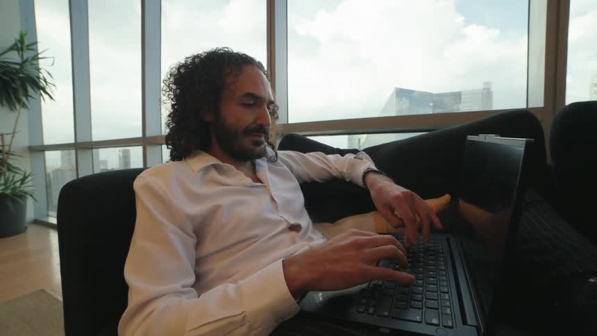 A relaxed man with curly hair works from home on his laptop, sitting comfortably on a couch in a modern high-rise apartment with a city view