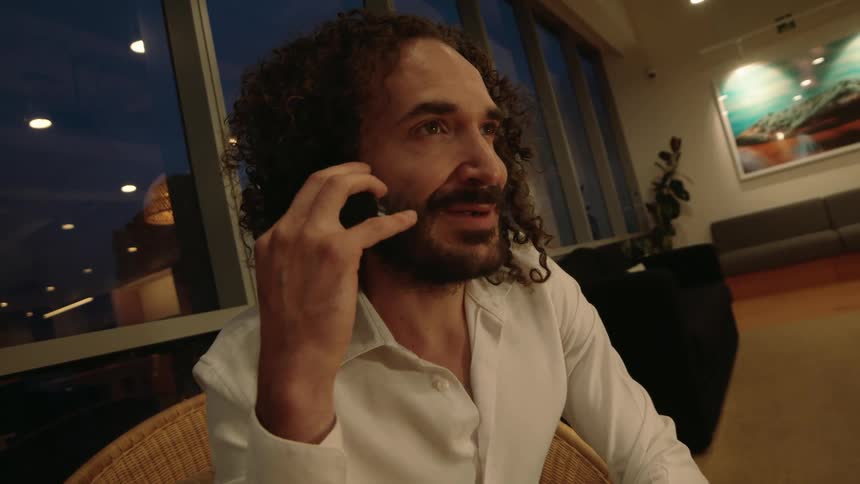 A handsome man with curly hair answers his smartphone and has a pleasant conversation in a modern lounge at night