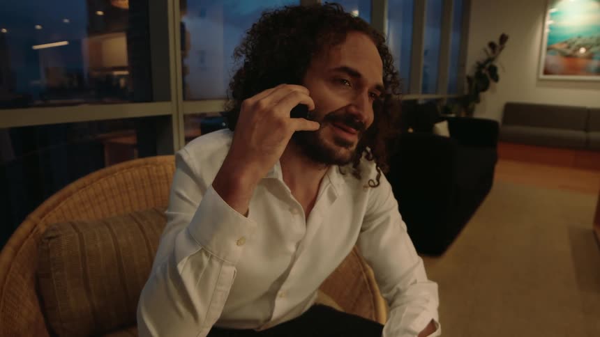 A handsome man with long curly hair and a beard has a pleasant conversation on his smartphone in a modern living room at night