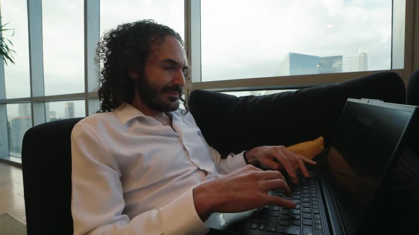 A relaxed professional man with curly hair works on his laptop from a sofa in a modern high-rise office