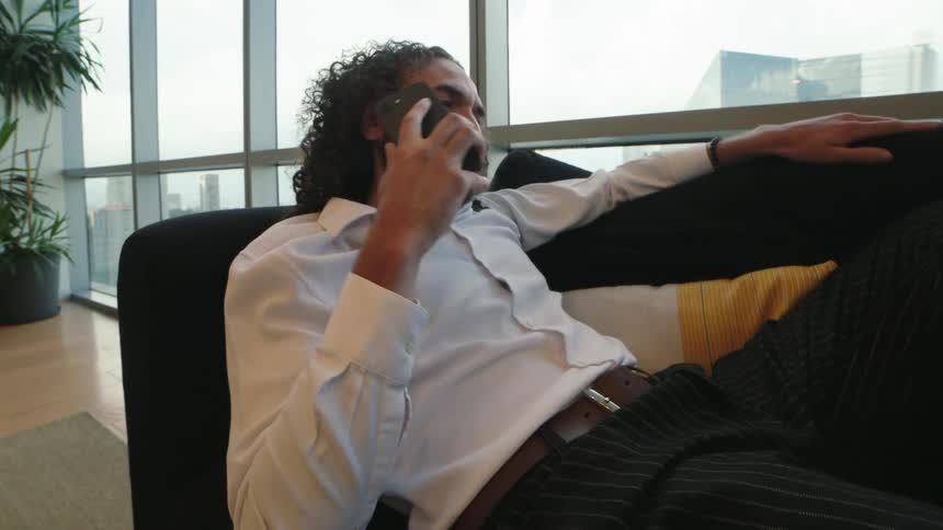 A relaxed businessman with long curly hair lounges on a couch in a modern high-rise office while taking a phone call