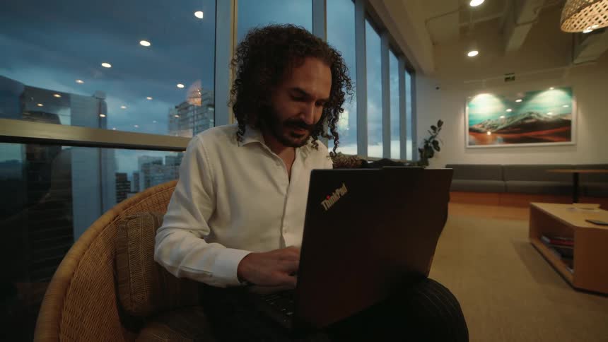 A focused businessman with curly hair types on his laptop in a modern high-rise office as dusk settles over the city