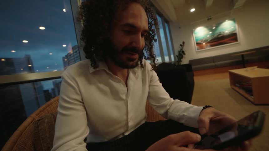 A stylish man with curly hair smiles while using his smartphone in a modern lounge overlooking the city at dusk