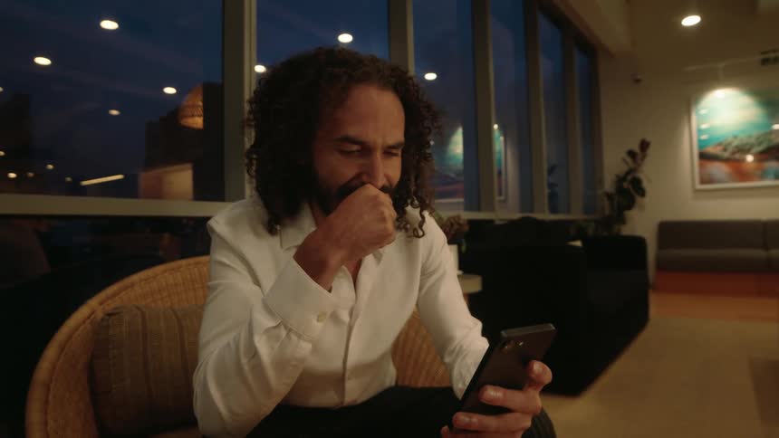 A thoughtful man with long curly hair scrolls on his smartphone while relaxing in a modern lounge in the evening