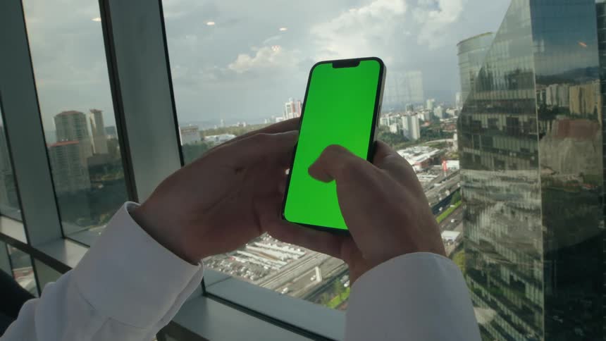 POV of a businessman scrolling on a green screen smartphone in a high-rise office with a cityscape view