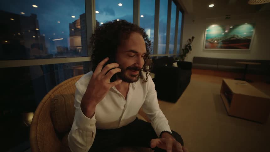 A businessman in a modern high-rise office smiles while taking a pleasant phone call in the evening