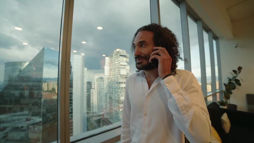 A successful businessman with curly hair smiles while talking on the phone in his modern high-rise office overlooking the city