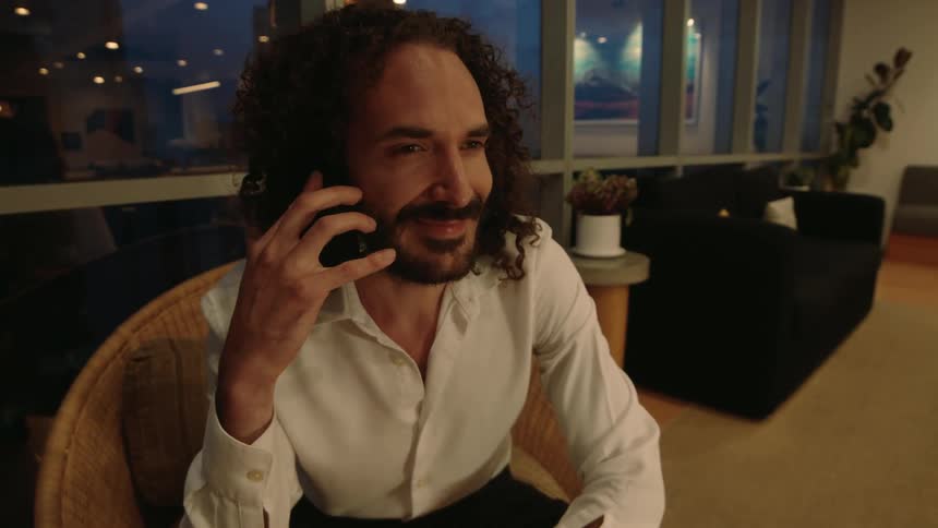 A man with curly hair smiles while talking on his smartphone in a modern lounge at night