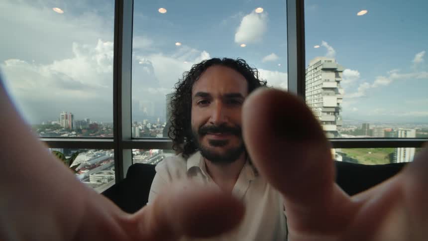A man with long curly hair smiles warmly in a first-person perspective as fingers playfully interact with the screen in front of a cityscape view
