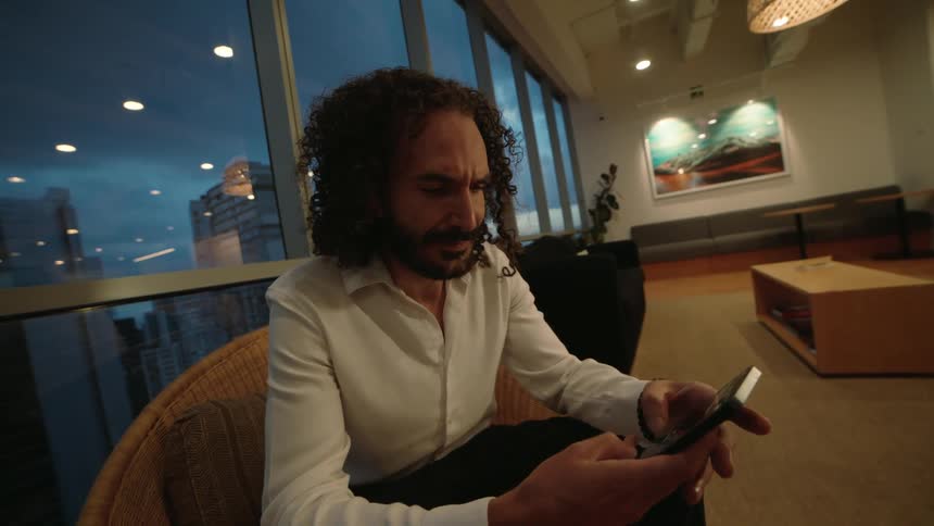 A businessman with long curly hair sits in a modern office lounge in the evening, intently focused on scrolling through his smartphone