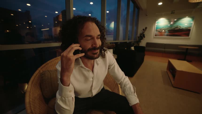 A professional man with curly hair sits in a comfortable chair while having a pleasant conversation on his phone in a modern office lounge at night
