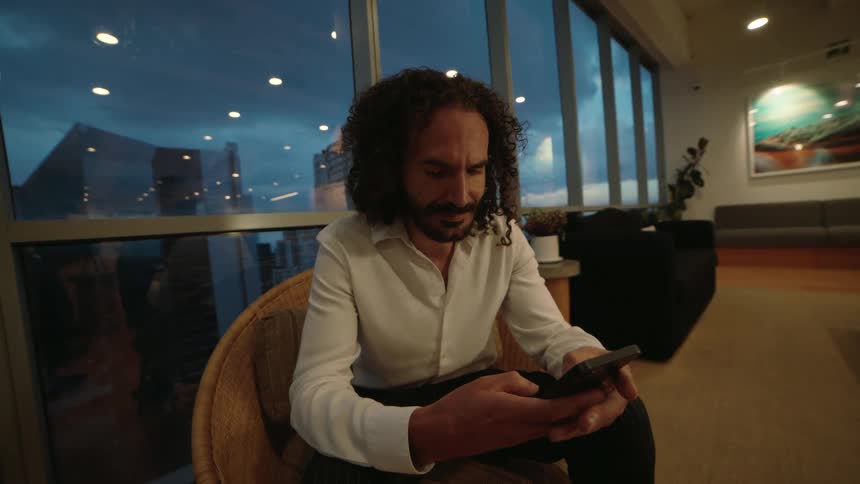 Stylish curly-haired man scrolls on his phone in a modern high-rise office lounge as evening falls over the city