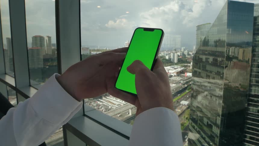 A businessman in a white shirt scrolls on a green screen smartphone in a modern high-rise office overlooking the city