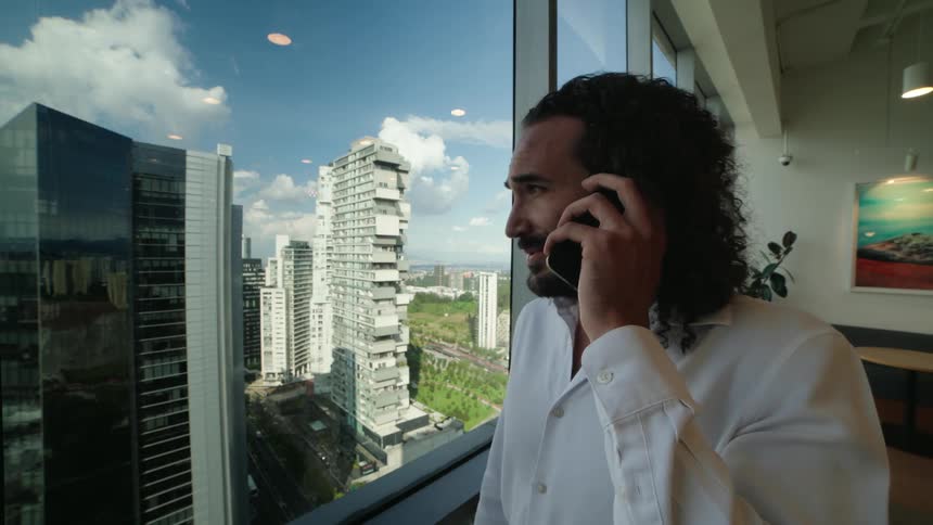 A handsome businessman with long hair talks on his phone while looking out the window of a modern high-rise office with a city view