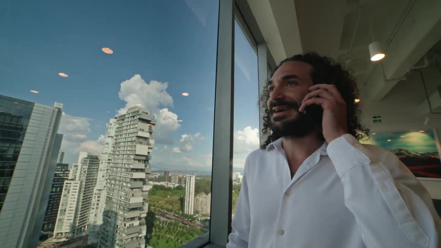 A happy businessman with curly hair smiles while talking on the phone in a high-rise office with a view of the cityscape