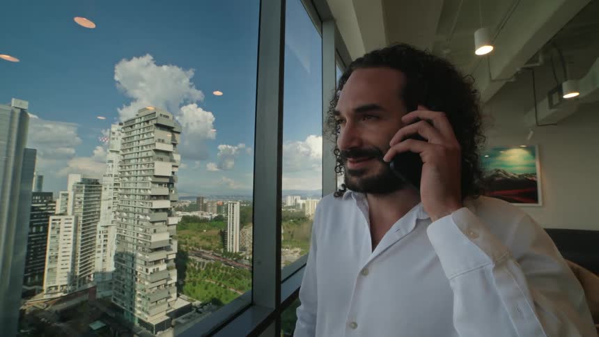A man with long hair and a beard smiles while making a phone call in a high-rise office overlooking the city skyline