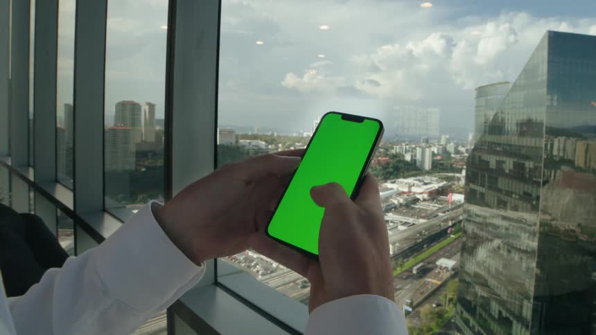 A businessman's hands scroll and tap on a green screen smartphone in a high-rise office with a city view