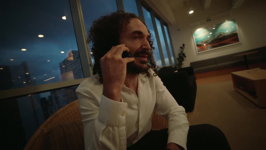 A businessman with long curly hair talks on his smartphone while sitting in a modern office lounge with a city view at night
