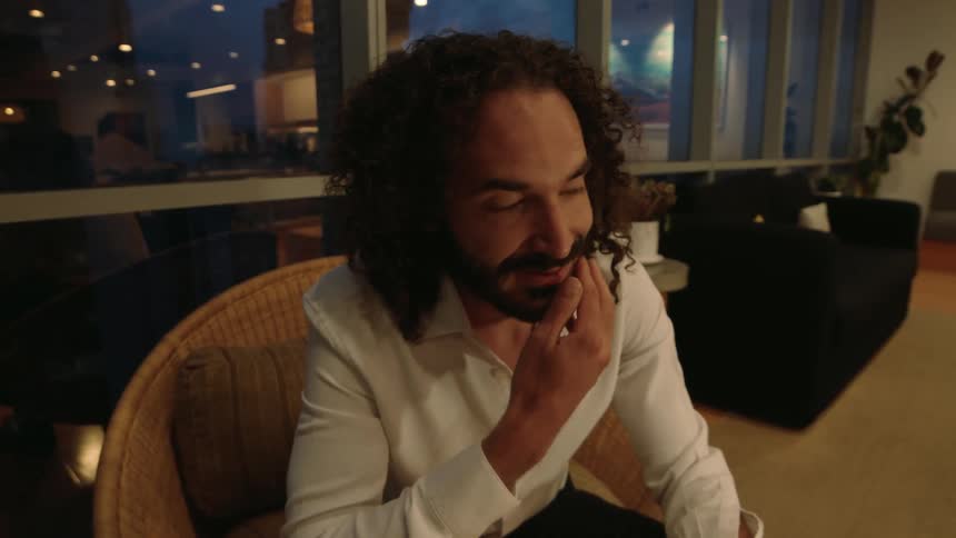 A thoughtful man with long curly hair and a beard sits in a chair in a modern apartment at dusk, gently stroking his chin