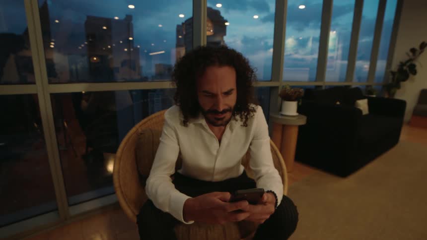 A stylish man with curly hair sits in a wicker chair in his modern high-rise apartment at night, focused on his smartphone