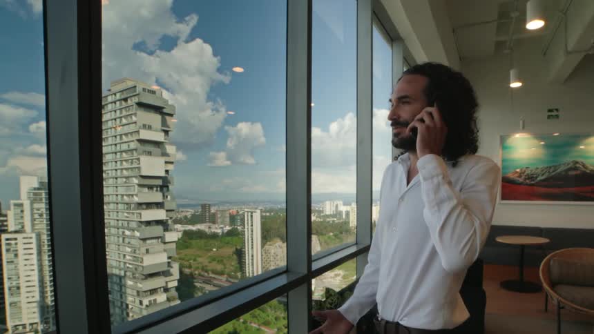 A successful businessman talks on the phone while looking out a large window with a panoramic view of the city