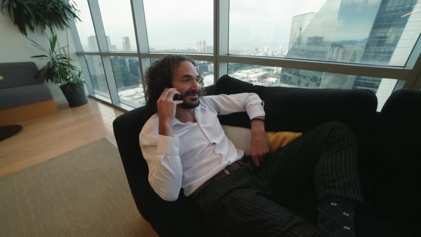 A stylish man with long curly hair relaxes on a sofa in a modern high-rise apartment and has a friendly conversation on his mobile phone