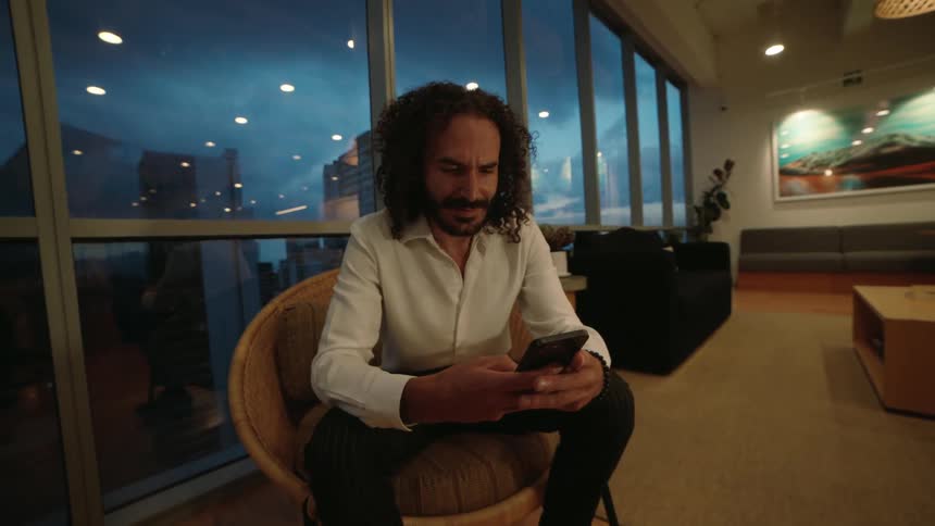 A stylish businessman with long curly hair uses his smartphone while sitting in a modern high-rise office lounge at dusk