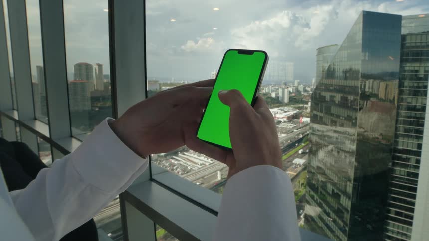 Businessman scrolls on a green screen smartphone in a high-rise office with a city view