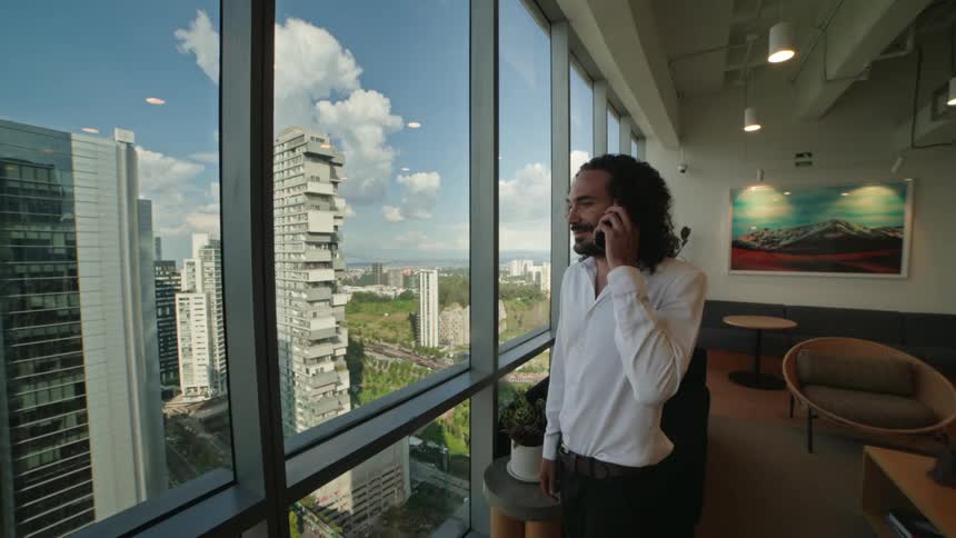 A handsome businessman with long hair talks on his phone as he walks to a large window in his modern high-rise office to enjoy the city view