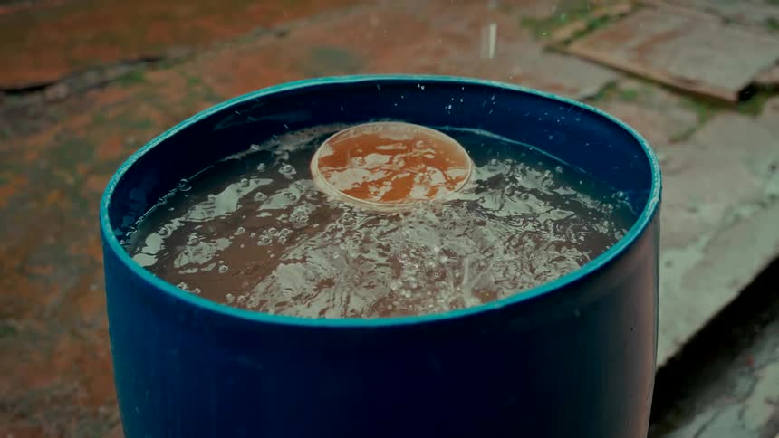 Raindrops fall in slow motion into a blue plastic barrel collecting rainwater outside